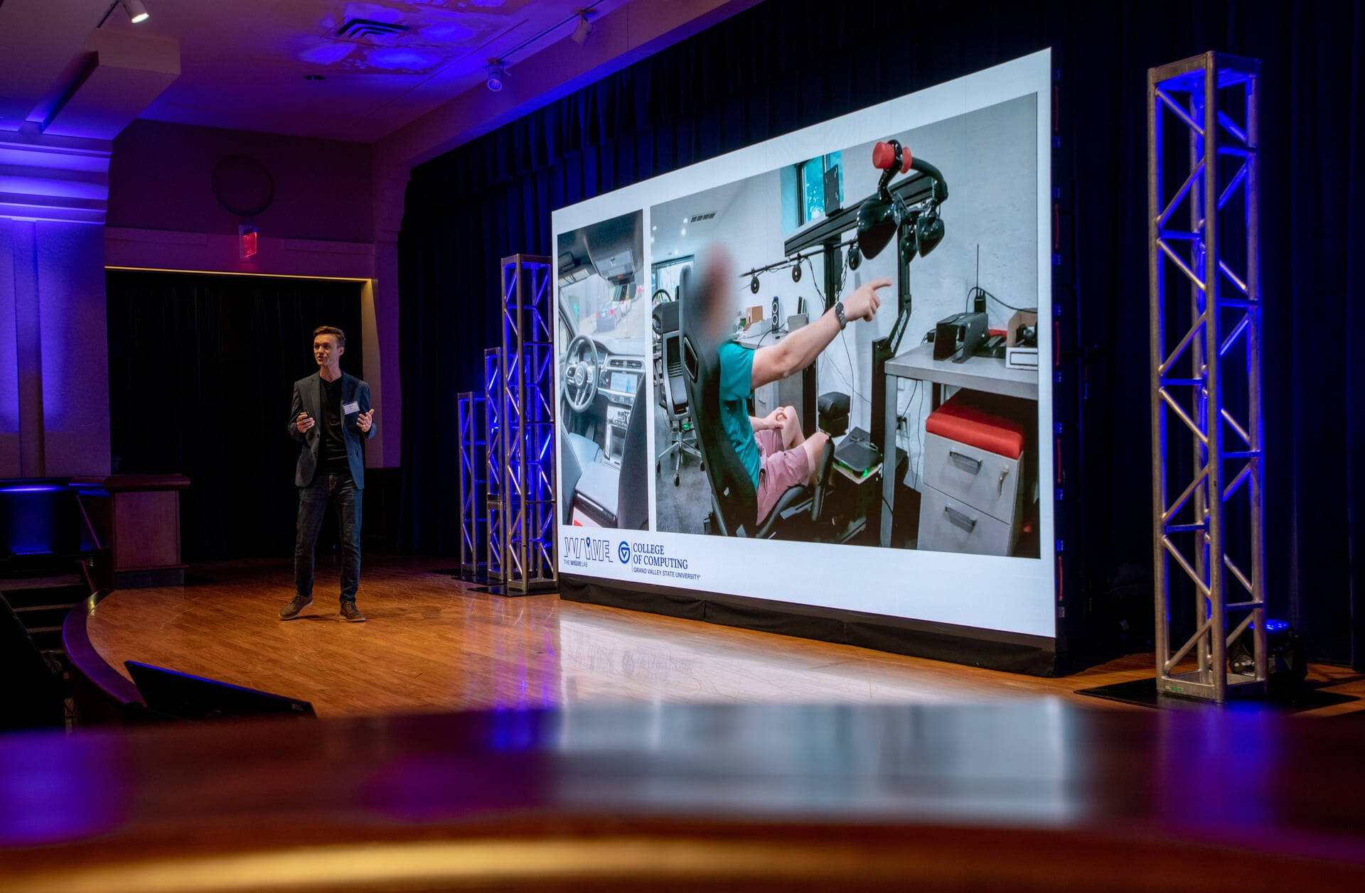 Paul Fink, assistant professor in the College of Computing, gives remarks on stage at Loosemore Auditorum, large screen showing adaptive vehicle behind him